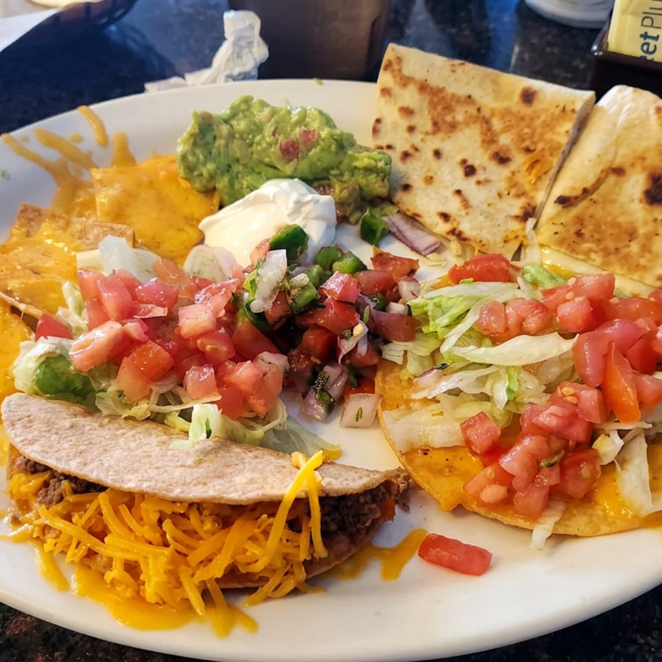 Mexican platter with a cheese-covered taco, tostadas, quesadillas, guacamole, sour cream, lettuce, and pico de gallo.