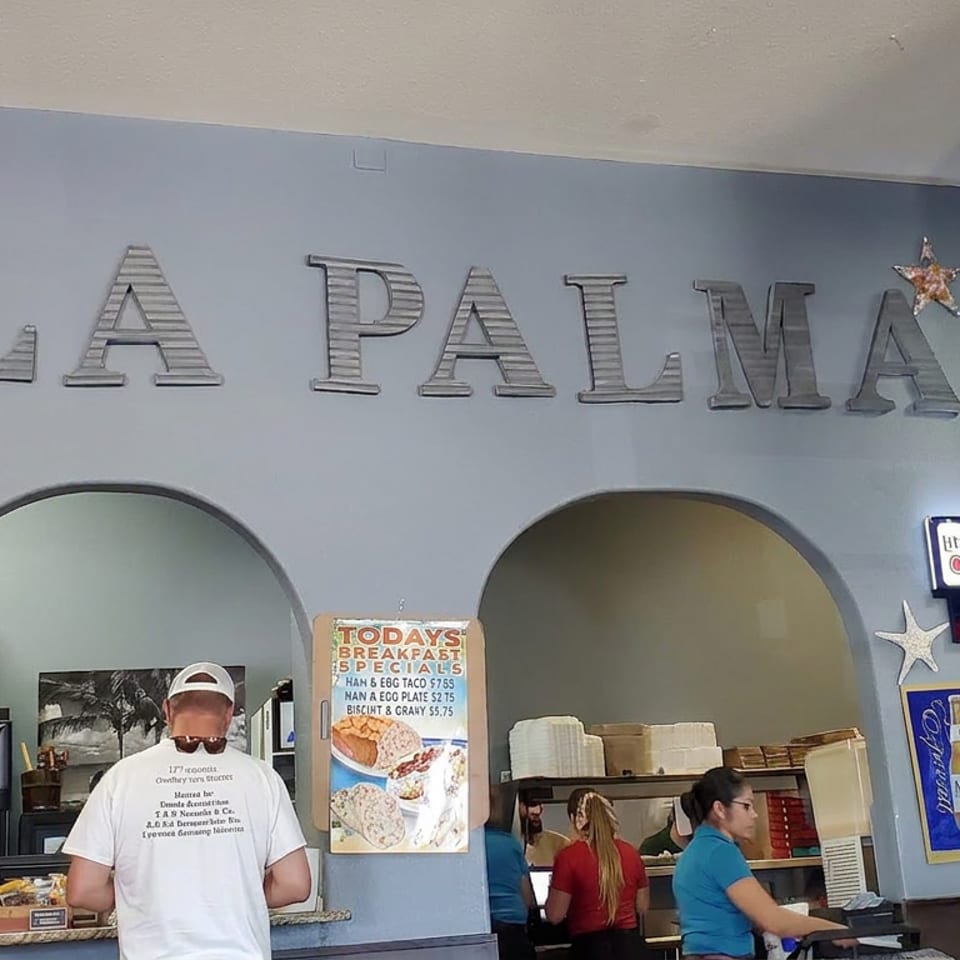La Palma restaurant interior with large wall lettering, arched service windows, staff behind the counter, and breakfast specials sign.