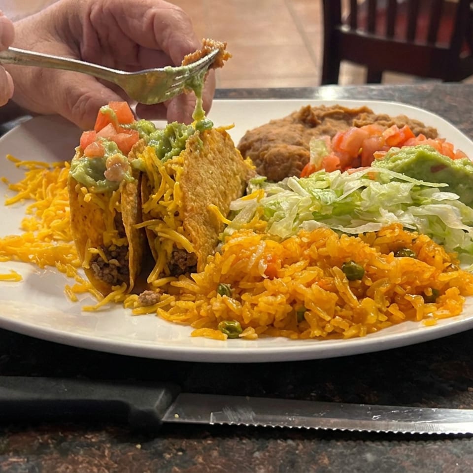 Plate of crispy beef tacos with shredded cheese, guacamole, lettuce, salsa, refried beans, and Mexican rice, while a fork lifts a bite.