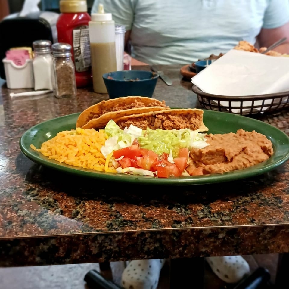 Plate of crispy tacos with Mexican rice, refried beans, guacamole, lettuce, and tomatoes on a restaurant table.
