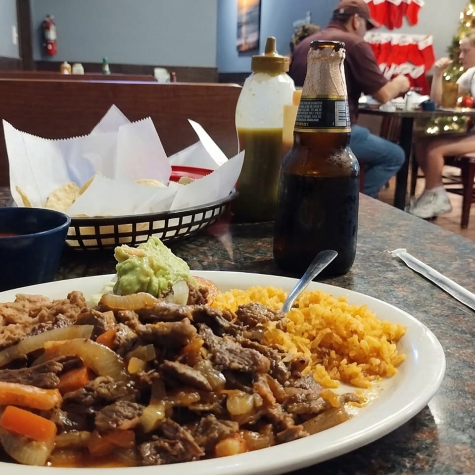 Restaurant table featuring carne asada with onions and peppers, refried beans, yellow rice, guacamole, tortillas, salsa, and a bottled beer.