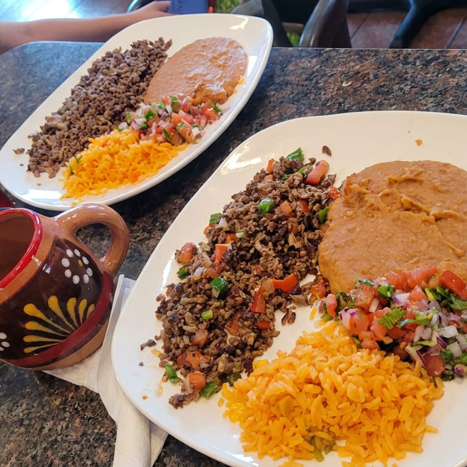Two plates of Mexican-style rice, seasoned ground beef, refried beans, and pico de gallo on a restaurant table.